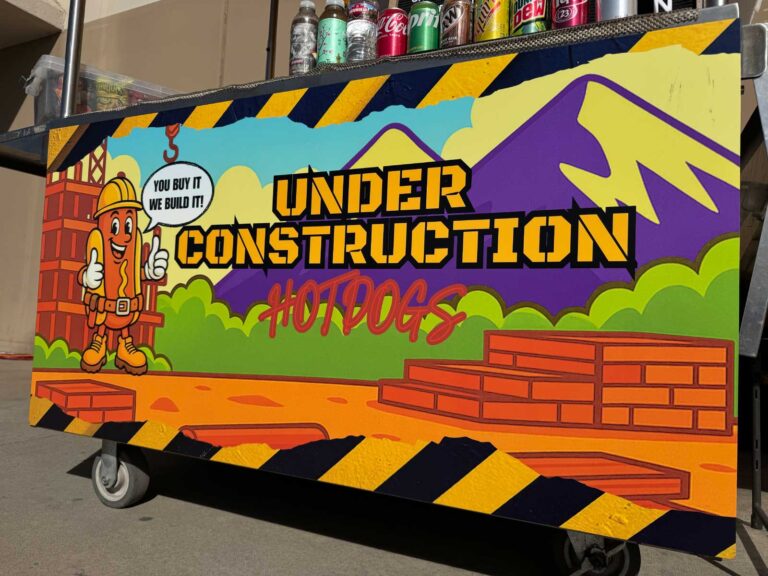 The Under Construction Hot Dogs cart, painted with a cartoon hot dog in a hard hat and the line You buy it! We build it!