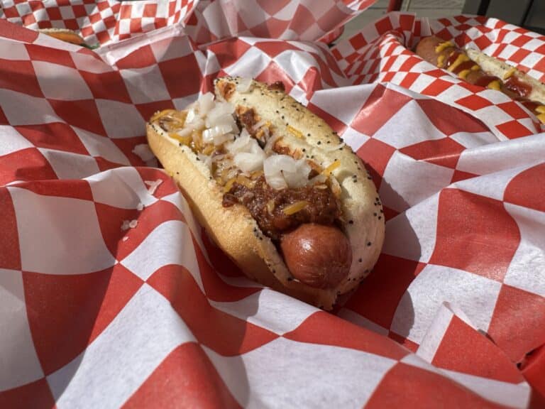 A chili cheese hot dog topped with chopped onions on red and white checkered paper at Under Construction Hot Dogs.