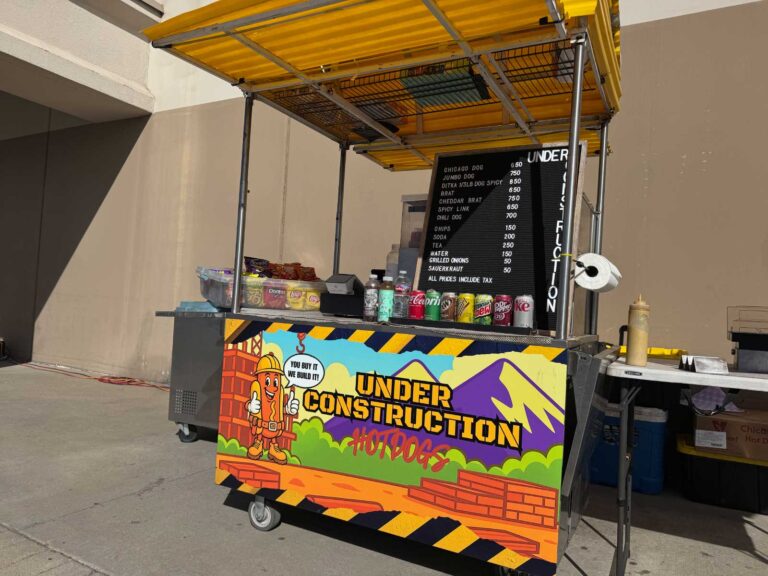 The Under Construction Hot Dogs cart set up outside with its menu board, snacks, and drinks.